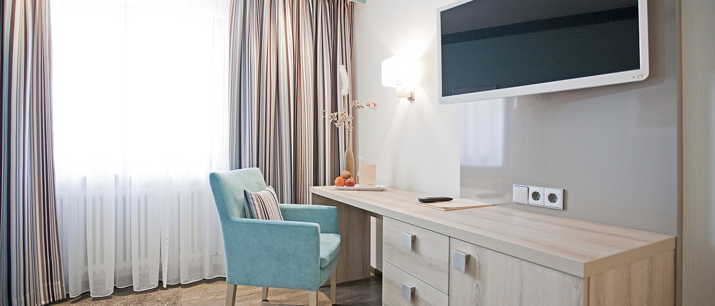 Modernes Einzelzimmer mit Schreibtisch, blauem Stuhl, Flachbildfernseher und gro&szlig;en Fenstern mit gestreiften Vorh&auml;ngen., &copy; Hotel Krone