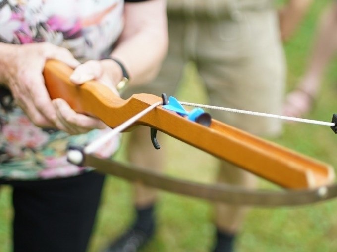 Nahaufnahme einer Person, die eine Armbrust mit einem blauen Pfeil im Freien hält. Im Hintergrund unscharf Gras und Beine sichtbar., © Cool-Tours StattReisen Nahaufnahme einer Person, die eine Armbrust mit einem blauen Pfeil im Freien hält. Im Hintergrund unscharf Gras und Beine sichtbar., © Cool-Tours StattReisen
