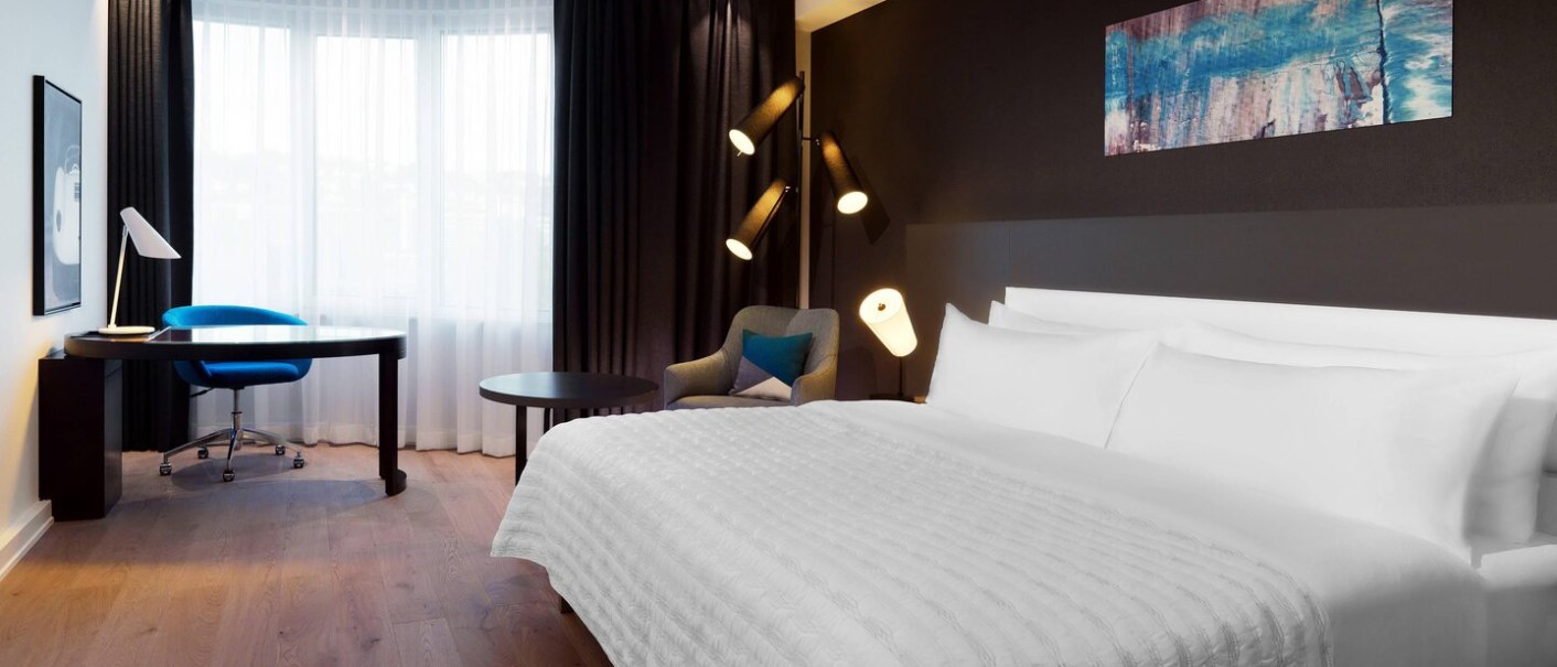 Modernes Hotelzimmer mit großem Bett, Schreibtisch, blauem Stuhl und Sessel. Helle Vorhänge und stilvolle Beleuchtung., © Hotel Le Méridien Modernes Hotelzimmer mit großem Bett, Schreibtisch, blauem Stuhl und Sessel. Helle Vorhänge und stilvolle Beleuchtung., © Hotel Le Méridien