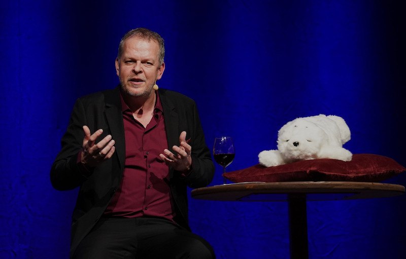 Ein Mann sitzt auf einer Bühne, spricht gestikulierend. Neben ihm ein Tisch mit einem Glas Wein und einem weißen Plüschtier auf einem Kissen., © Theaterhaus Stuttgart e.V.
