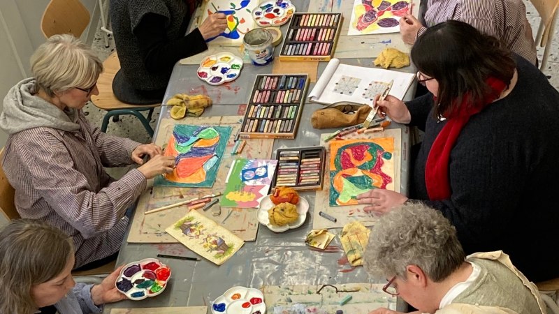 Teilnehmer eines Kunstworkshops sitzen an einem Tisch und arbeiten mit Farben und Pastellkreiden an bunten Kunstwerken., &copy; Adolf H&ouml;lzel Stiftung