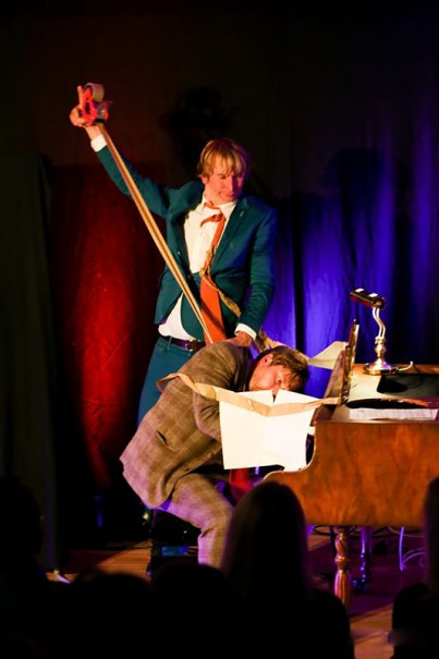 Zwei M&auml;nner f&uuml;hren eine kom&ouml;diantische Szene auf. Einer h&auml;lt ein gro&szlig;es Gummiband, w&auml;hrend der andere seinen Kopf in eine Kiste auf einem Tisch steckt., &copy; Theaterhaus Stuttgart e.V.