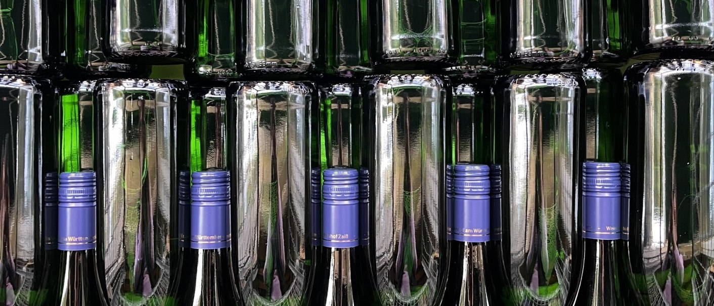 Stacked green wine bottles with blue caps on a shelf. The bottles reflect the light, creating a symmetrical pattern., &copy; Weinhof Zai&szlig;