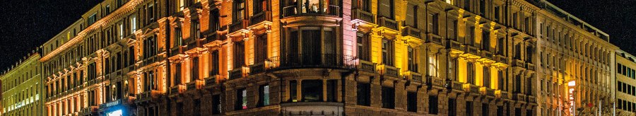 A large, illuminated building at night with stores on the first floor and people on the street., © Schauspielbühnen in Stuttgart A large, illuminated building at night with stores on the first floor and people on the street., © Schauspielbühnen in Stuttgart