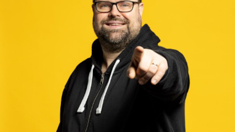 Mann mit Brille und Bart zeigt lächelnd auf die Kamera. Er trägt einen schwarzen Hoodie. Der Hintergrund ist leuchtend gelb., © Theaterhaus Stuttgart e.V.