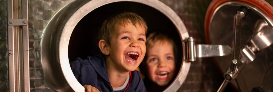 Two children laugh happily out of the round opening of a large metal container., © Weingut Zaiß