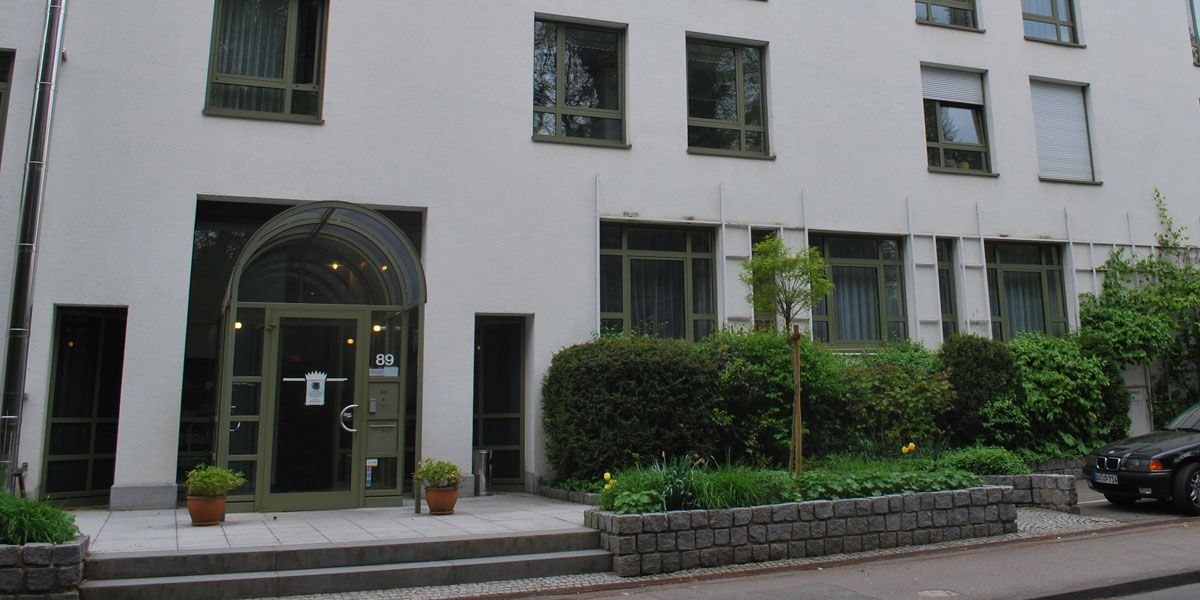 A modern building entrance with a glass roof and a green door. There are potted plants in front of the entrance. A car is parked on the right., &copy; Christk&ouml;nigshaus