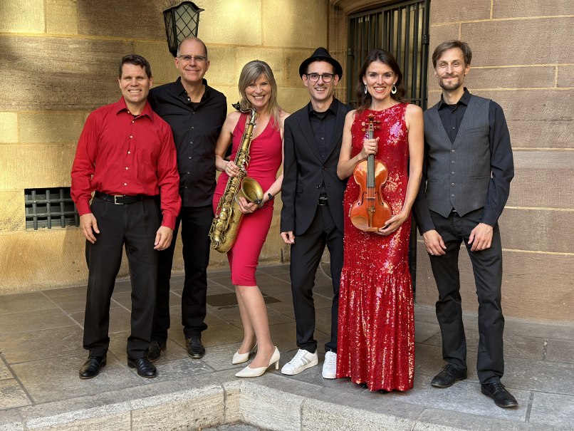 Sechs Personen in festlicher Kleidung posieren lächelnd. Zwei halten Musikinstrumente: ein Saxophon und eine Geige. Hintergrund aus Steinmauern., © Friedrichsbau Varieté