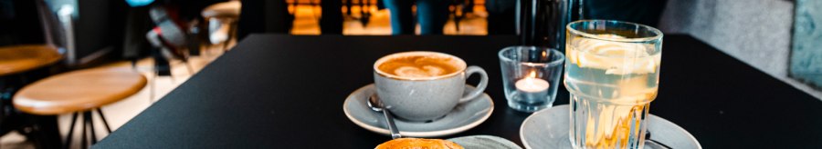 A table in a café with a cinnamon bun, cappuccino and lemon water. Blurred people can be seen in the background., © SMG Stuttgart Marketing GmbH - Sarah Schmid