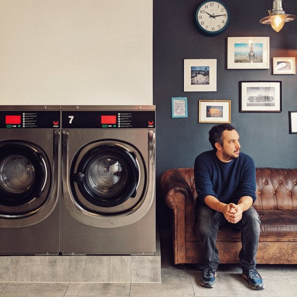 Ein Mann sitzt auf einem Ledersofa in einem Waschsalon neben Waschmaschinen. An der Wand h&auml;ngen Bilder und eine Uhr., &copy; Veranstalter