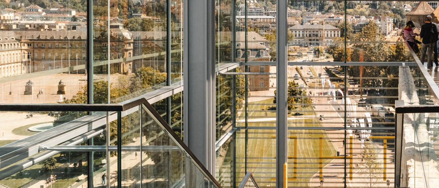 Blick durch moderne Glasarchitektur auf den Schlossplatz. Im Hintergrund historische Gebäude und Menschen. Klare Linien und Reflexionen dominieren das Bild., © Stuttgart-Marketing GmbH, Sarah Schmid Blick durch moderne Glasarchitektur auf den Schlossplatz. Im Hintergrund historische Gebäude und Menschen. Klare Linien und Reflexionen dominieren das Bild., © Stuttgart-Marketing GmbH, Sarah Schmid