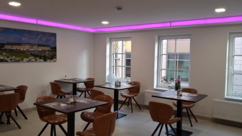 Moderner Loungebereich mit lila Beleuchtung, Tischen und Stühlen. An der Wand hängt ein großes Bild, Fenster lassen Tageslicht herein., © TOMAS