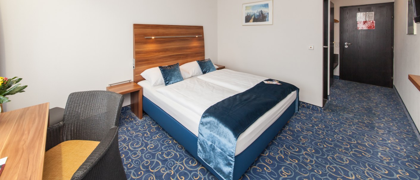 Ein Hotelzimmer mit Doppelbett, blauem Teppich und Holzmöbeln. Ein Schreibtisch mit Stuhl und ein Bild an der Wand sind sichtbar., © Novum Management GmbH Ein Hotelzimmer mit Doppelbett, blauem Teppich und Holzmöbeln. Ein Schreibtisch mit Stuhl und ein Bild an der Wand sind sichtbar., © Novum Management GmbH