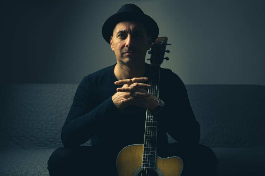 Ein Mann mit schwarzem Hut sitzt auf einem Sofa und hält eine Akustikgitarre. Die Beleuchtung ist gedämpft und erzeugt eine ruhige Atmosphäre., © Foto: Tom Lumen