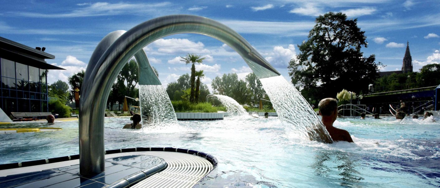 Ein Außenpool mit Wasserfontänen und Palmen, umgeben von Bäumen und einem Gebäude. Menschen genießen das Wasser unter blauem Himmel., © Stuttgarter Bäder Ein Außenpool mit Wasserfontänen und Palmen, umgeben von Bäumen und einem Gebäude. Menschen genießen das Wasser unter blauem Himmel., © Stuttgarter Bäder