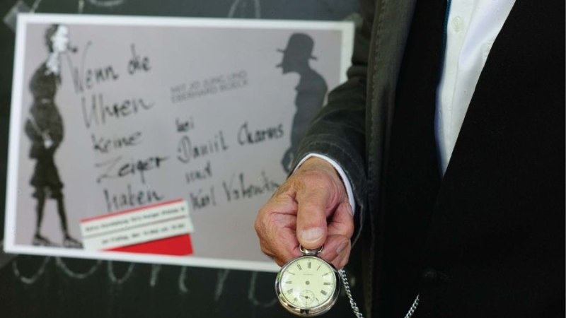 Ein Mann in Anzug h&auml;lt eine Taschenuhr vor einem Plakat mit dem Titel 'Wenn die Uhren keine Zeiger haben'., &copy; Jim Zimmermann
