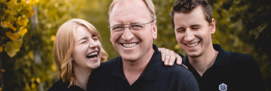 Three people in black clothing are laughing heartily. They are standing in front of a blurred green background. One shirt reads 'Schwarz Weingut'., © Weingut Schwarz