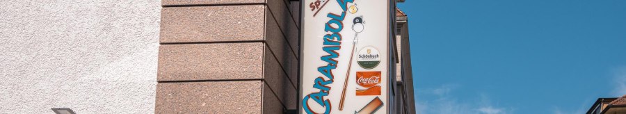 Ein Schild mit der Aufschrift 'Carambolage' hängt an einer Gebäudewand. Der Himmel ist klar und blau., © SMG, Sarah Schmid Ein Schild mit der Aufschrift 'Carambolage' hängt an einer Gebäudewand. Der Himmel ist klar und blau., © SMG, Sarah Schmid