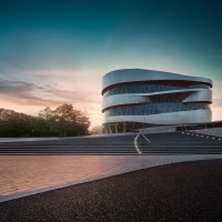Das Mercedes-Benz Museum in Stuttgart bei Sonnenuntergang. Moderne Architektur mit geschwungenen Linien, umgeben von Bäumen und beleuchteten Stufen., © Mercedes-Benz Heritage GmbH