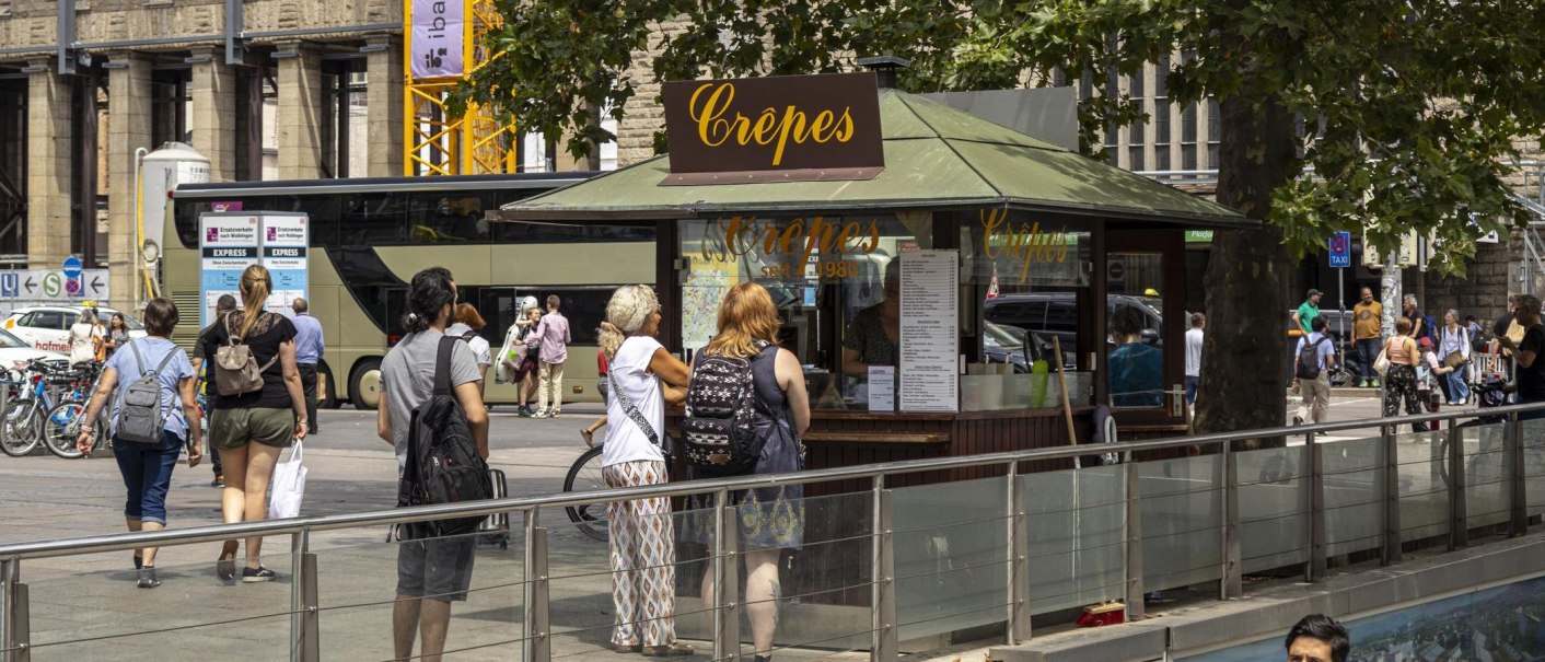 Ein Crêpes-Stand am Stuttgarter Hauptbahnhof mit mehreren Menschen, die anstehen. Im Hintergrund sind Fahrräder und ein Bus zu sehen., © SMG, Sarah Schmid Ein Crêpes-Stand am Stuttgarter Hauptbahnhof mit mehreren Menschen, die anstehen. Im Hintergrund sind Fahrräder und ein Bus zu sehen., © SMG, Sarah Schmid