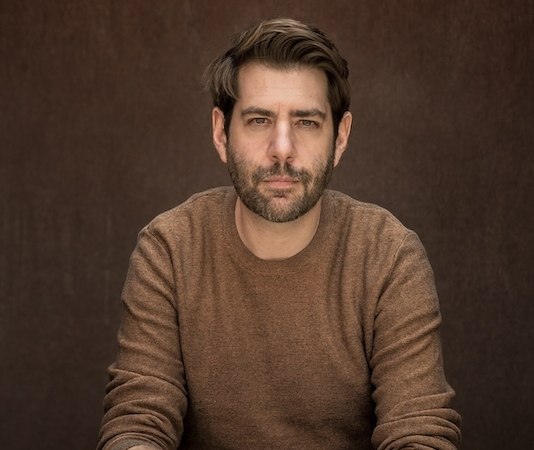 A man in a brown sweater sits with his hands clasped in front of a brown background and looks seriously into the camera., &copy; Renitenztheater Stuttgart e.V.