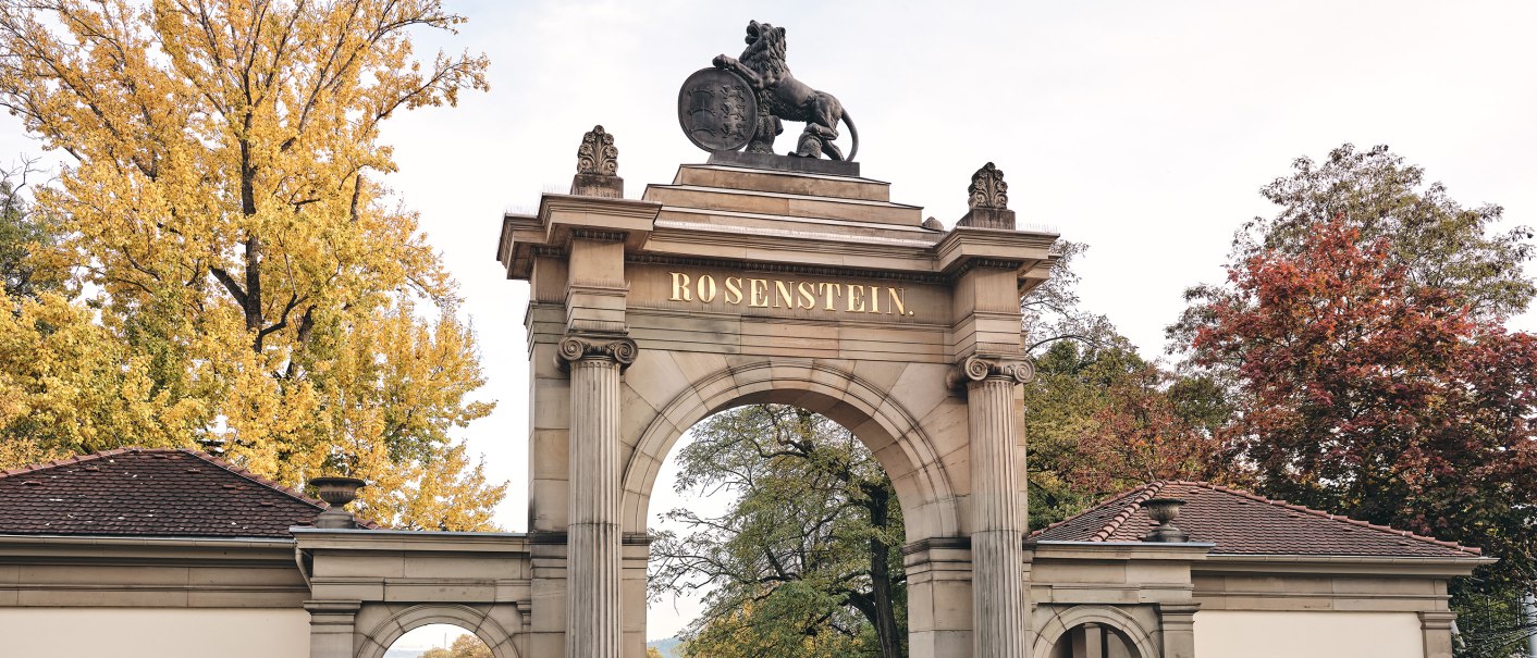 Historic gate with 'Rosenstein' lettering, flanked by buildings. Autumnal trees in the background, two people walking., © TMBW, Christoph Düpper