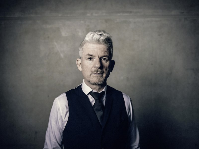 A man with gray hair wears a suit and stands in front of a gray wall. He looks seriously into the camera., &copy; Theaterhaus Stuttgart e.V.