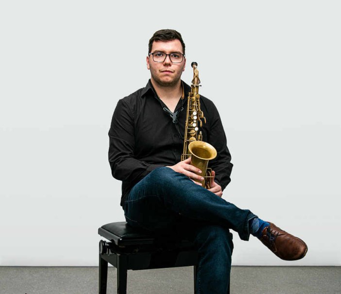 Ein Mann in schwarzem Hemd und Jeans sitzt mit einem Saxophon auf einem Stuhl. Der Hintergrund ist neutral., © Lars Roth