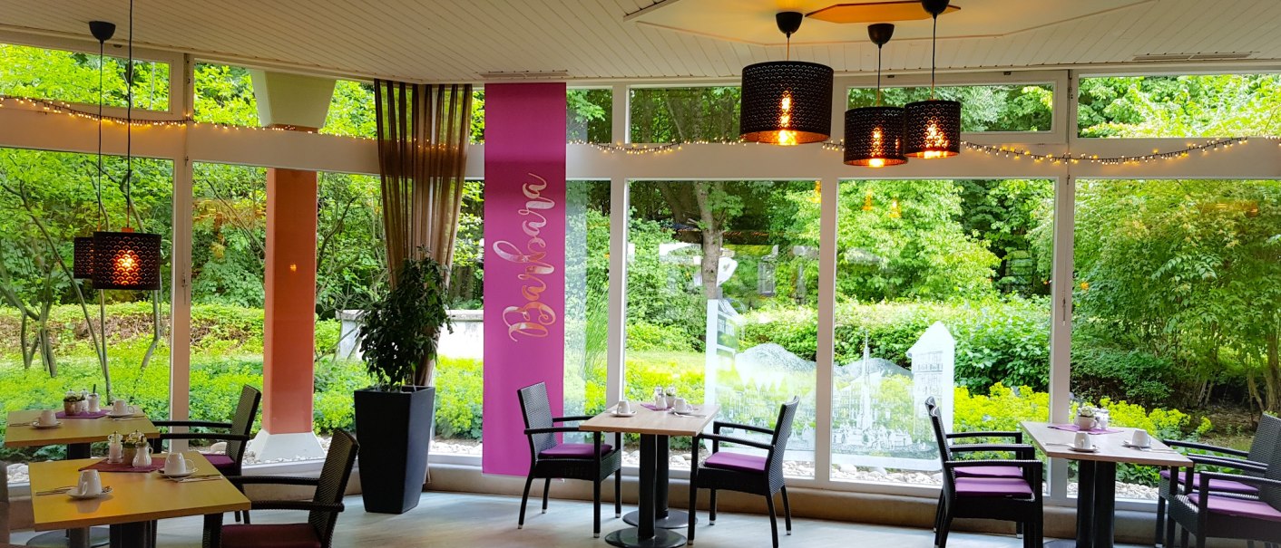 Ein gemütliches Café mit großen Fenstern, die auf einen grünen Garten blicken. Tische sind mit Geschirr gedeckt, und Lampen hängen von der Decke., © ©hotel-graf-eberhard.de