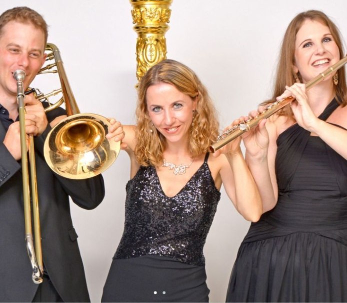 Drei Musiker in Abendkleidung posieren mit Posaune und Flöte vor einem goldenen Dekor. Sie lächeln und wirken fröhlich., © Trio Glissando Stuttgart