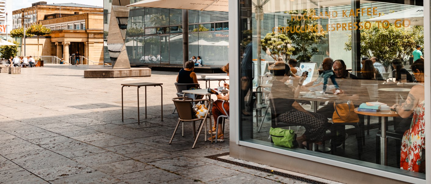 Außenansicht eines Cafés mit Glasfront, Menschen sitzen an Tischen, moderne Architektur im Hintergrund., © SMG, Sarah Schmid Außenansicht eines Cafés mit Glasfront, Menschen sitzen an Tischen, moderne Architektur im Hintergrund., © SMG, Sarah Schmid