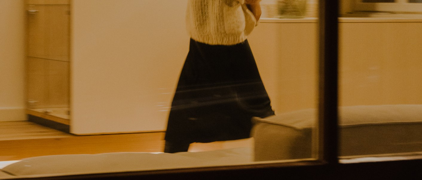 Eine Frau in einem hellen Raum, durch ein Fenster fotografiert. Sie tr&auml;gt einen wei&szlig;en Pullover und einen langen schwarzen Rock. Die Beleuchtung ist warm., &copy; Peter Oliver Wolff / DAS EDITH