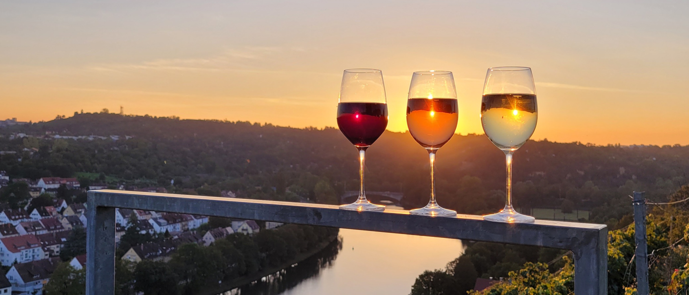 Drei Weingläser mit Rot-, Rosé- und Weißwein stehen auf einem Geländer. Im Hintergrund ein Sonnenuntergang über einer Flusslandschaft., © Weinbau Glock und Sohn GbR