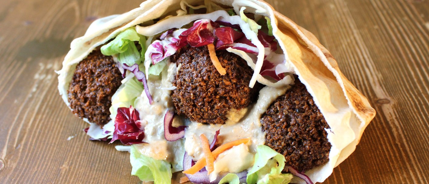A falafel wrap with crispy falafel balls, fresh salad and sauce in a flatbread on a wooden table., © Vegi Stuttgart