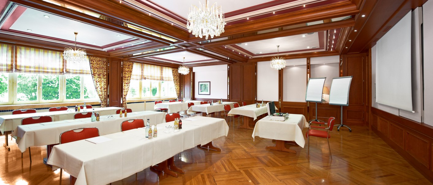 Eleganter Seminarraum mit gedeckten Tischen, roten Stühlen, Flipcharts und Kronleuchtern. Große Fenster lassen viel Licht herein., © Hirsch Hotel