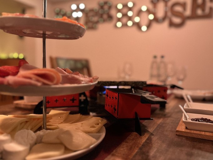 A table with raclette grill, cheese, cold cuts and side dishes. Blurred decoration and glasses in the background., © Cool-Tours StattReisen A table with raclette grill, cheese, cold cuts and side dishes. Blurred decoration and glasses in the background., © Cool-Tours StattReisen
