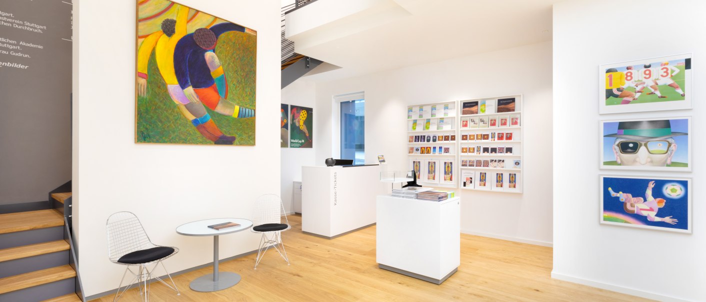 A modern art gallery with colorful paintings, light-colored wooden floors, and a reception desk. Two chairs and a table are located to the left of a staircase., © Freundeskreis Fritz Genkinger e.V. A modern art gallery with colorful paintings, light-colored wooden floors, and a reception desk. Two chairs and a table are located to the left of a staircase., © Freundeskreis Fritz Genkinger e.V.