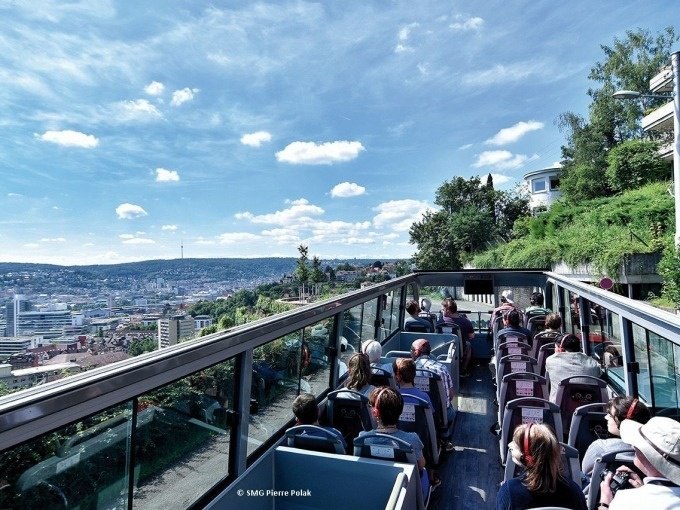 Tourists sit in an open double-decker bus and enjoy the view of the city of Stuttgart under a blue sky with clouds., © Willms Touristik Tourists sit in an open double-decker bus and enjoy the view of the city of Stuttgart under a blue sky with clouds., © Willms Touristik